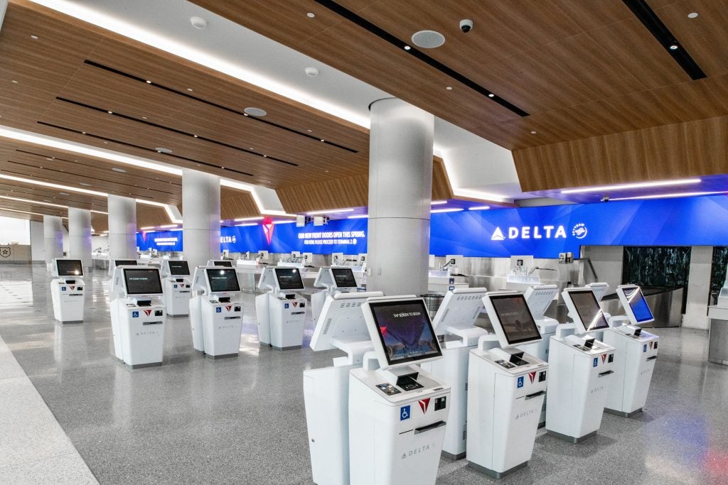 An airport check in area with many self-check in stations consisting of one screen atop a white podium. Delta's logo can be seen on screens in the background of the area.