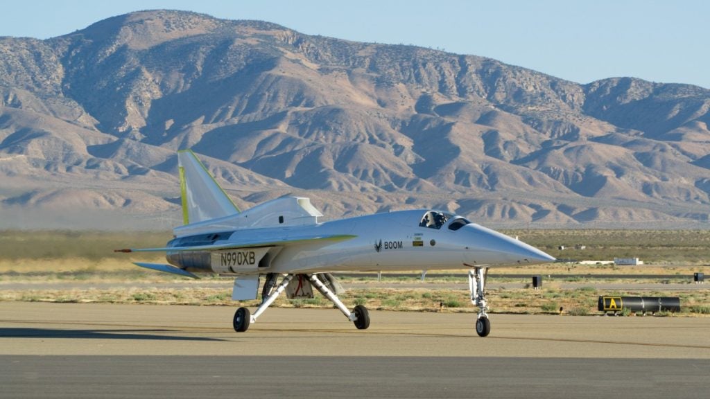 The XB-1, a small, grey supersonic jet sits on the tarmac at the Mojave air and space port with mountains and desert in the background.